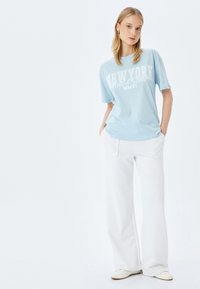 Lichtblauwe katoenen T-shirt met de opdruk "NEW YORK United States MMVI", gecombineerd met witte wijde broek en lichte schoenen. Eenvoudige, casual look.