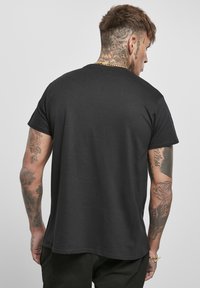 T-shirt noir à manches courtes avec un col rond, texture lisse et un ourlet légèrement courbé. Tatouages visibles sur les bras.