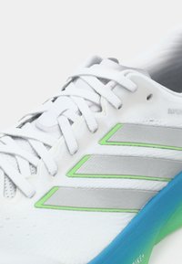 Λευκό αθλητικό παπούτσι με ασημένιες και πράσινες λεπτομέρειες, με υφασμένο επάνω μέρος, παραδοσιακά κορδόνια και σόλα με μπλε διαβαθμίσεις.