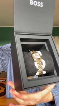Relógio dourado com um mostrador quadrado, pulseira ligada e marcações brancas, exibido sobre uma almofada escura numa caixa preta. Logótipo da Boss visível.