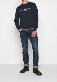 Mörkblå crewneck sweatshirt med vit text "BELSTAFF", kombinerad med mörkblå jeans och svarta läderankelstövlar med kraftig sula.