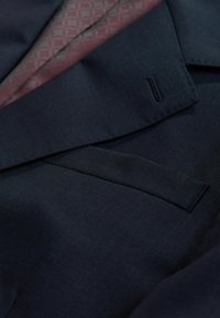Dunkelblauer Blazer mit strukturierter Oberfläche, dezentem Taschen-Detail und kontrastierendem Innenfutter mit geometrischem Muster in Bordeaux.