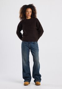 Brauner strukturierter Pullover, entspannte Passform, runder Halsauschnitt; kombiniert mit lockeren blauen Jeans und braunen Schuhen. Minimalistisches Styling, neutrale Farben.