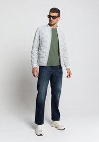 Lichtgrijze gewatteerde bomberjack over een donkergroen T-shirt, gecombineerd met donkerblauwe jeans en witte sneakers met grijze accenten. Zonnebril op.