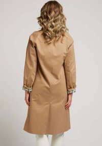 Manteau trench beige avec des manches longues et une coupe ajustée. Présente un détail de poignet à carreaux. Texture lisse avec un léger éclat.