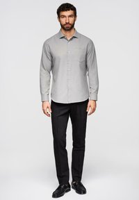 Homme debout, portant une chemise à manches longues gris clair à boutons, un pantalon noir et des chaussures habillées noires, sur un fond blanc uni.