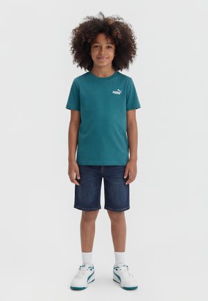 Bambino con capelli ricci che indossa una maglietta teal Puma, pantaloncini di jeans, calzini bianchi e scarpe da ginnastica bianche, in piedi davanti a uno sfondo plaino.