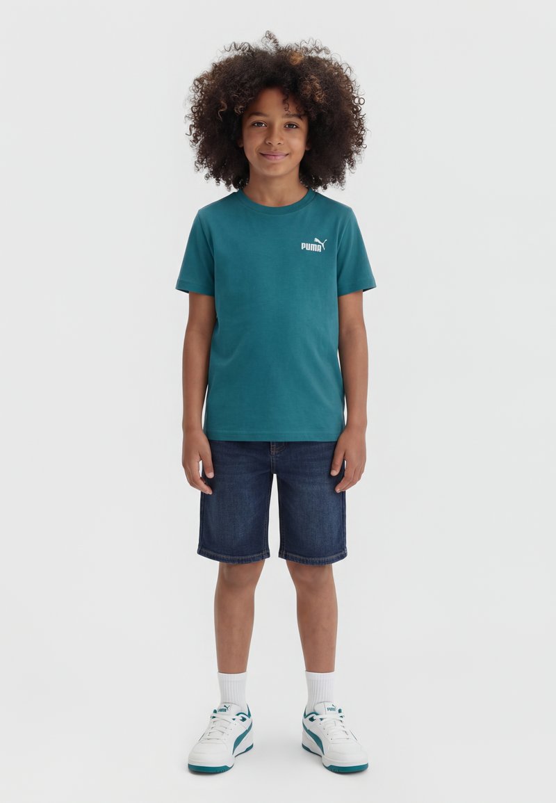 Дете с къдрави коси, облечено в тюркоазена тениска Puma, дънкови къси панталони, бели чорапи и бели спортни обувки, стои пред едноцветен фон.