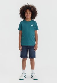 Παιδί με κατσαρά μαλλιά, που φοράει πετρόλ κοντομάνικη μπλούζα Puma, τζιν σορτς, άσπρες κάλτσες και άσπρα αθλητικά παπούτσια, στέκεται μπροστά σε απλό φόντο.