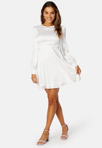 Abito bianco in satin a maniche lunghe con corpino aderente e gonna svasata. Presenta polsini delle maniche arricciati e una superficie liscia. Abbinato a tacchi color lavanda.