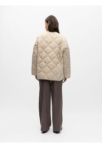 Γυναίκα, ορατή από πίσω, φοράει μπεζ καπιτονέ μπουφάν και φαρδιά καφέ παντελόνια με χαλαρή εφαρμογή, μπροστά σε λευκό απλό φόντο.