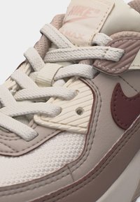 Κοντινή λήψη στο μπροστινό μέρος και τα κορδόνια του αθλητικού παπουτσιού Nike Air Max σε μπεζ και καφέ αποχρώσεις, με ορατές υφές υφάσματος και λεπτομέρειες λογοτύπου.