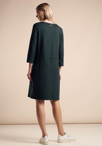 Robe-sweat vert foncé avec des manches trois-quarts, silhouette décontractée, détail de couture au dos et accent logo près du col.