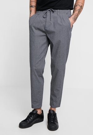 Mężczyzna ubrany w szare spodnie z troczkami i czarne trampki, ręce w kieszeniach, widoczne tatuaże na przedramionach, stoi na jednolitym tle.