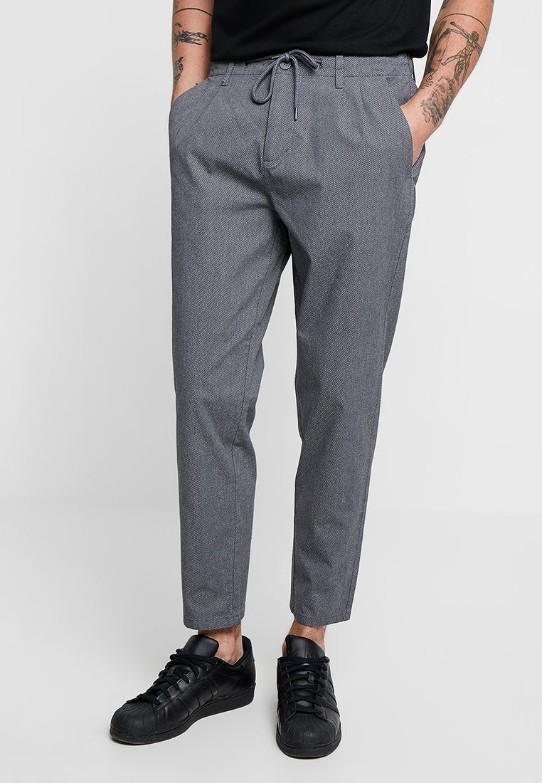 Homme portant un pantalon gris à cordon de serrage et des baskets noires, les mains dans les poches, tatouages visibles sur les avant-bras, debout devant un fond uni.