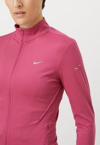 Lähikuva henkilöstä, joka on pukeutunut vaaleanpunaiseen vetoketjulliseen urheilu takkiin, jossa on pieni heijastava Nike-logo rinnassa ja heijastavia yksityiskohtia hihassa.