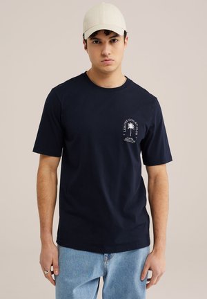 Młody mężczyzna w beżowej czapce, granatowej koszulce z białym logo palmy i napisem oraz jasnoniebieskich dżinsach, stojący na jednolitym tle.