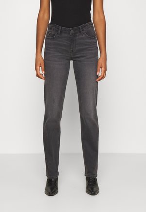 Persoon die vervaagde zwarte jeans met rechte pijpen, een zwart aansluitend shirt en zwarte leren puntige schoenen draagt, staand tegen een effen lichte achtergrond.