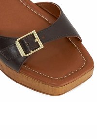 Bruin leren slip-on sandaal met een gouden gesp, bruine binnenzool en houten zool. Beschikt over witte stiksels en een vierkante toel ontwerp.