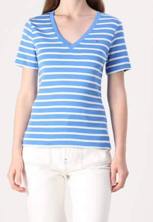 Femme portant un t-shirt à manches courtes rayé bleu et blanc à col en V et un pantalon blanc, montrée du cou aux hanches.
