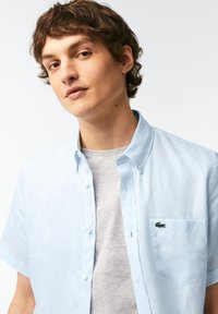 Camicia a maniche corte azzurro chiaro con bottoni e taschino sul petto, con logo ricamato verde. Indossata sopra una t-shirt grigia chiara.