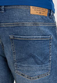Farkut sinisellä pesulla, jossa on nahkainen tägi, jossa lukee "DENIM DRIVEN SINCE 1989", ja kontrastiväriset keltaiset tikit takataskussa.