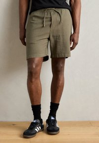 Olivegrønne shorts i midt-lår længde med elastisk talje og snøre. Stoffet har en tekstureret overflade. Bæres med sorte sneakers og sokker.