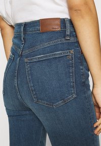 Person med blå högmidjade jeans med bakfickor, en lädermärke, och en vit skjorta som är instoppad synlig vid midjan.