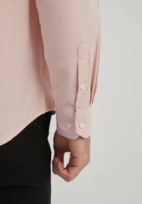 Chemise rose clair avec un motif texturé, dotée d'un poignet boutonné et d'une légère courbe à l'ourlet, associée à un pantalon noir.