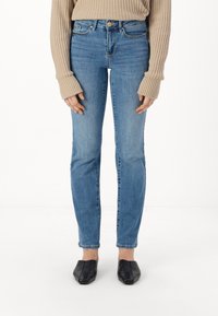 Pantalones vaqueros de mezclilla azul de tiro medio con un ajuste slim, que cuentan con un diseño de cinco bolsillos y un solo botón frontal. Combinados con un suéter de color beige de canalé.