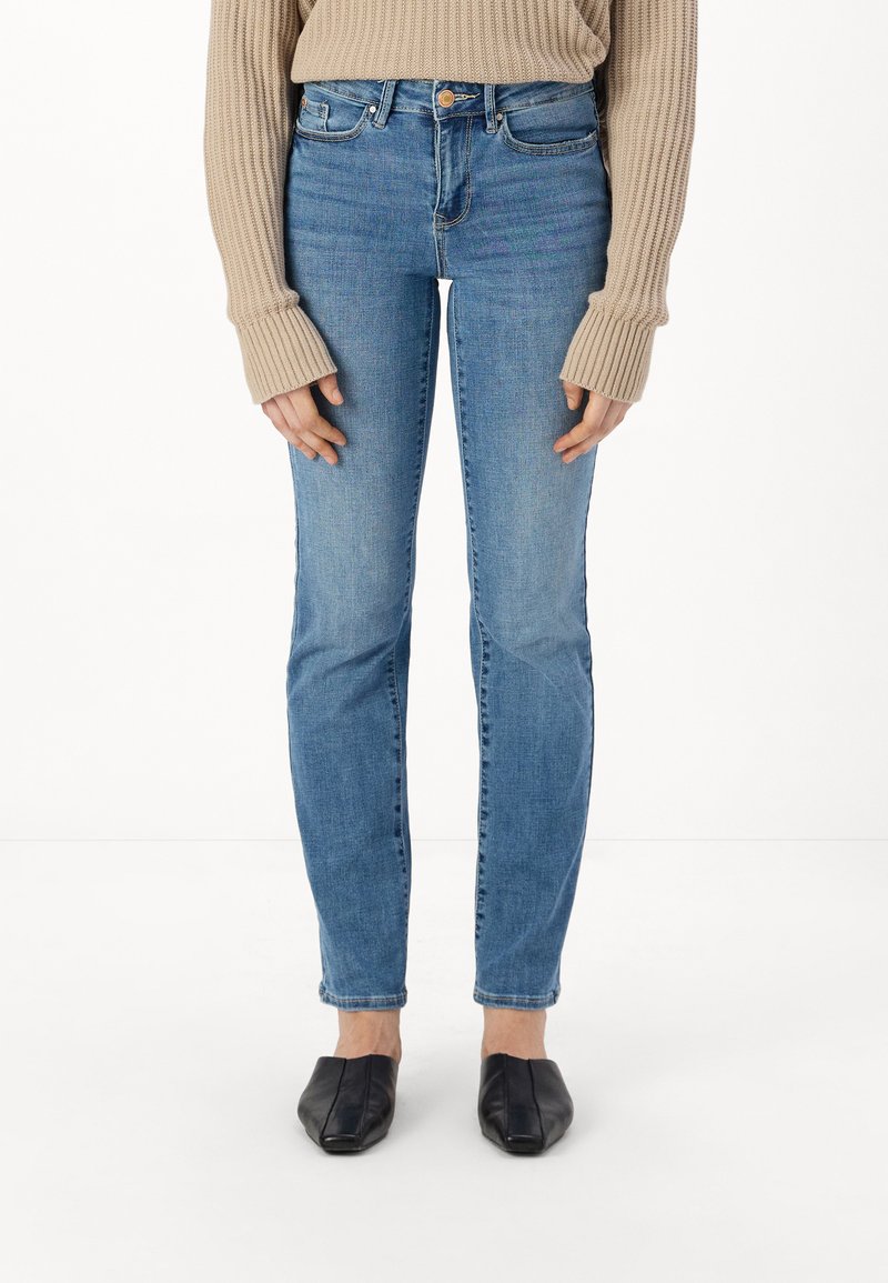 Pantalones vaqueros de mezclilla azul de tiro medio con un ajuste slim, que cuentan con un diseño de cinco bolsillos y un solo botón frontal. Combinados con un suéter de color beige de canalé.