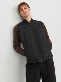 Gilet rembourré noir avec des coutures verticales, col haut et fermeture éclair frontale, porté sur une chemise à manches longues marron. Texture lisse et mate.