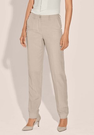 Vrouw draagt beige rechte broekspijpen en beige spitse hoge hakken, staand tegen een neutrale achtergrond.