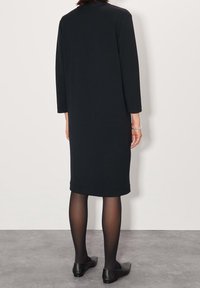 Černé šaty ke kolenům s dlouhými rukávy a jednoduchým kulatým výstřihem. Hladká textura, padají přímo z ramen. Nosené s černými balerínami.