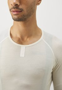 Helles beige, tailliertes Hemd mit rundem Halsausschnitt, glatter Textur und einem kleinen Markenetikett am Kragen. Verfügt über Raglanärmel.