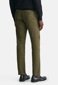 Olivgrüne Hose mit taillierter Passform, aus glattem Material und mit Gesäßtaschen mit Knopfverschluss, kombiniert mit schwarzen Mokassins.