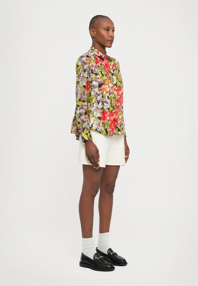 Bluză florală cu mânecă lungă în nuanțe strălucitoare de roșu, verde și mov, asortată cu pantaloni scurți albi și mocasini negri cu accente aurii.