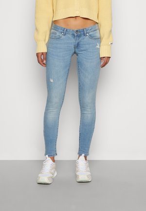Femme portant un jean skinny bleu clair avec une légère usure, un pull jaune court et des baskets blanches, debout sur un sol gris.