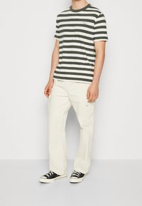 Hombre de pie vistiendo una camisa de manga corta a rayas verdes y blancas, pantalones cargo color crema y zapatillas negras y blancas contra un fondo plano.