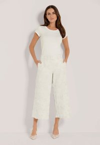 Witte fitted t-shirt gecombineerd met lichtgekleurde, gestructureerde broek met wijde pijpen, voorzien van bloemenborduursels, voorkruiken en een kortere lengte, afgemaakt met nude hakken.