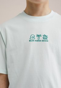 Svétlo modra bombažna majica z zelenim vezenim logotipom, ki prikazuje drevo palm in sonce, postavljena na sredini področja prsnega koša.