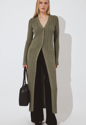 Ženska nosi dolgo olivno zeleno rebrasto jopico z gumbi preko črnih hlač, drži črno torbico, na belem ozadju.