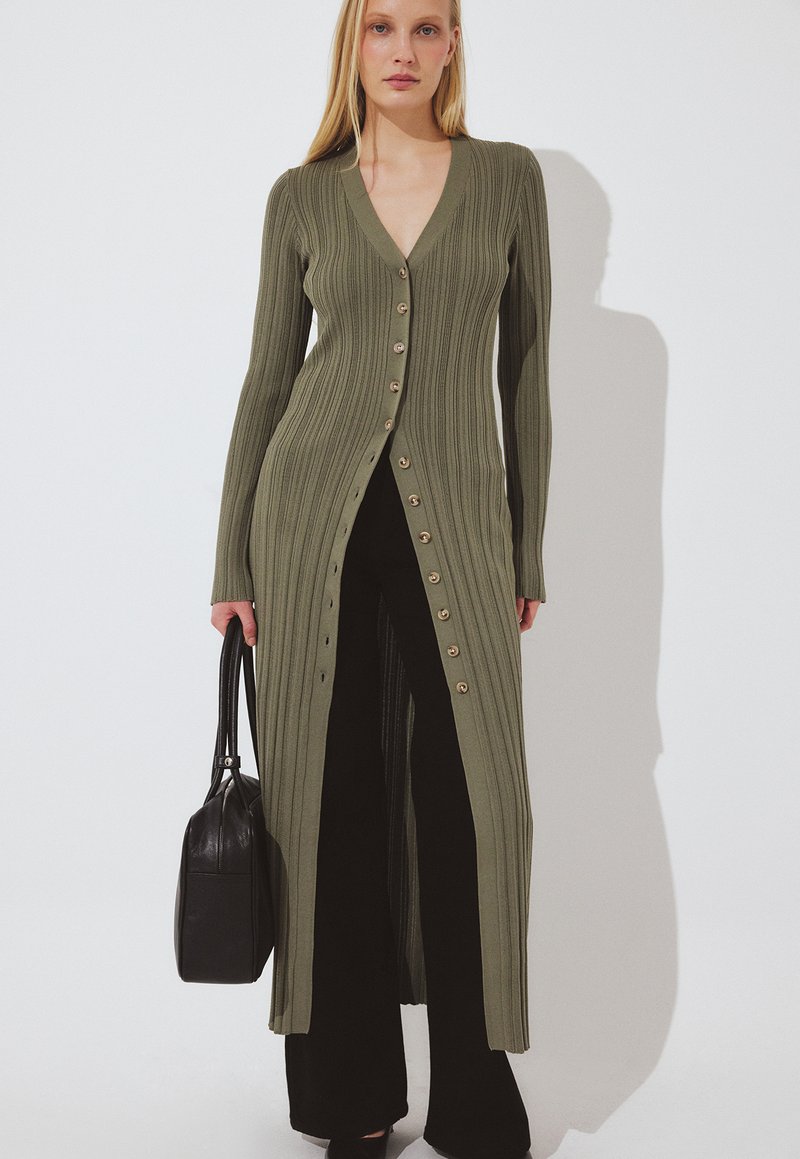 Moteris dėvi ilgą alyvuogių žalios spalvos ribinį megztinį su sagomis ant juodų kelnių, laiko juodą rankinę, fone baltas fonas.