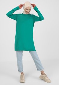 Haut vert à manches longues avec col montant, en tissu lisse, associé à un jean bleu clair avec revers et des baskets beiges.