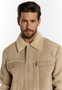 Beige Wildlederjacke mit einem Lammfellkragen, zwei Vordertaschen und ausgefransten Nähten, ausgestattet mit einem lässigen Schnitt und glatter Textur.