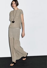 La modella cammina con grazia indossando un abito a righe con tessuto fluido; porta una lunga collana e sandali neri su uno sfondo minimalista.