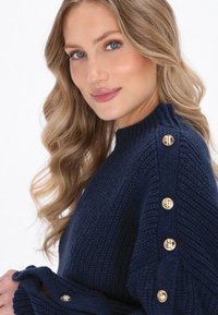 Pulover tricotat bleu marin cu textură canalată, având nasturi decorativi aurii pe umăr și un guler înalt.