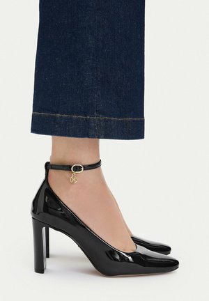 Kvinde iført sorte laklæder høje hæle med ankelsnore og guldfarvet vedhæng, kombineret med mørkeblå cropped denim bukser.