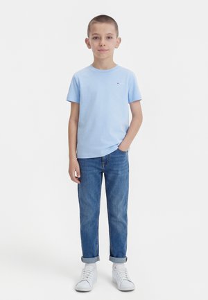 Αγόρι που στέκεται απέναντι κοιτάζοντας μπροστά, φορώντας ανοιχτό γαλάζιο κοντομάνικο μπλουζάκι, τζιν με ρεβέρ, λευκές κάλτσες και λευκά αθλητικά παπούτσια, με ένα χέρι στην τσέπη.