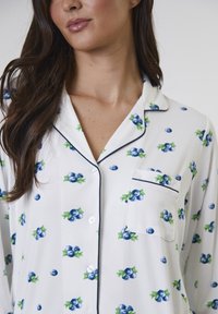 Weißes Schlafanzugoberteil mit blauem und grünem Heidelbeerblütenmuster, ausgestattet mit einer Tasche und dunkelblauem Paspel an den Rändern. Mit Knopfverschluss.